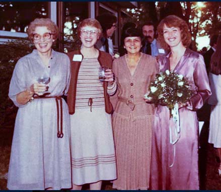 Four women in formal dresses smile towards the camera in a candid, outdoor wedding photo.