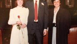 Wedding picture of Peter Marshall and his wife Roberta. Peter is wearing a dark suit with a red tie. Roberta is wearing a white, long-sleeved dress and holding a single red rose. They are standing next to Justice Abrahamson, who is wearing her judicial robes.
