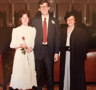 Wedding picture of Peter Marshall and his wife Roberta. Peter is wearing a dark suit with a red tie. Roberta is wearing a white, long-sleeved dress and holding a single red rose. They are standing next to Justice Abrahamson, who is wearing her judicial robes.