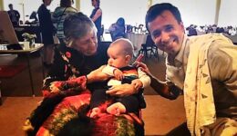 Shirley Abrahamson, seated, and holding a baby as Eli Best crouches next to her and smiles at the camera