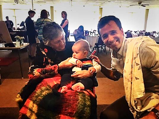 eli-best Shirley Abrahamson, seated, and holding a baby as Eli Best crouches next to her and smiles at the camera