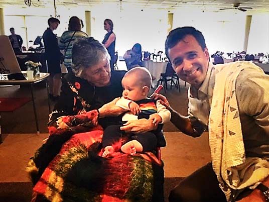 Shirley Abrahamson, seated, and holding a baby as Eli Best crouches next to her and smiles at the camera